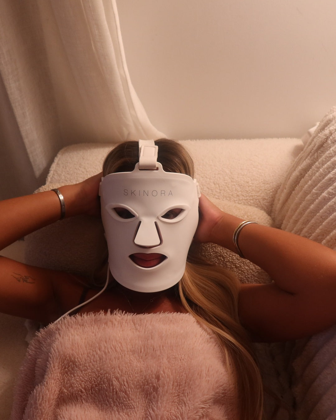 Masque LED Skinora porté éteint, mains derrière la tête, allongée sous un plaid